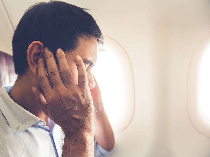 A man sits in an airplane seat next to a window, covering his ears with both hands, possibly due to discomfort or noise. The interior of the aircraft is softly lit, and the window shows a bright sky outside.