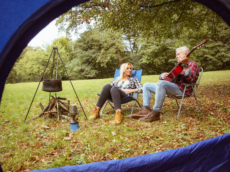 Two people sit in folding chairs outside a tent, surrounded by a grassy area and trees. One plays a guitar while the other listens, smiling. A tripod with a pot and a camp stove are nearby, suggesting they are camping.