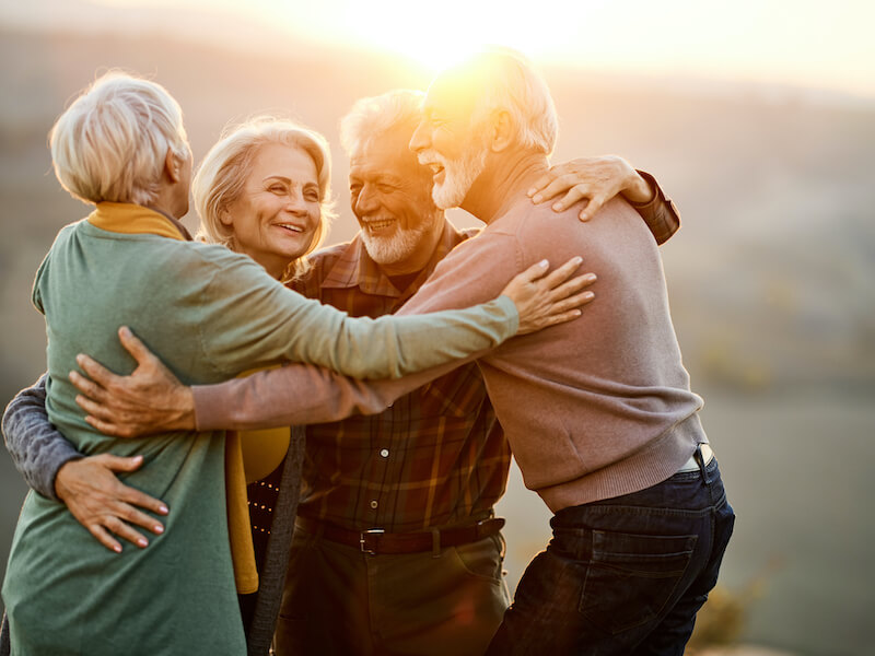 Four elderly friends embrace and smile joyfully in a sunlit outdoor setting. They appear relaxed and happy, enjoying each others company against a blurred natural background. The sunlight creates a warm and welcoming atmosphere.