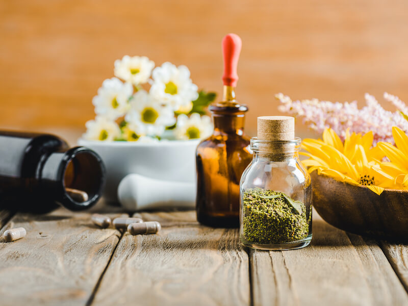 A rustic wooden table displays small glass bottles with corks, one filled with green herbs, and a brown bottle with a dropper. Nearby are capsules, a bowl of yellow flowers, and a mortar with white flowers, suggesting a natural remedy setting.