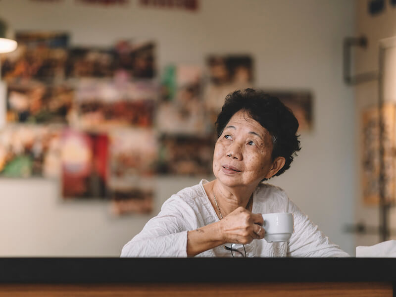 A woman, wearing a white top and glasses on a chain, sits at a table holding a white cup. She looks thoughtfully to the side. The background is a blurred wall with various photos. The atmosphere is calm and cozy.