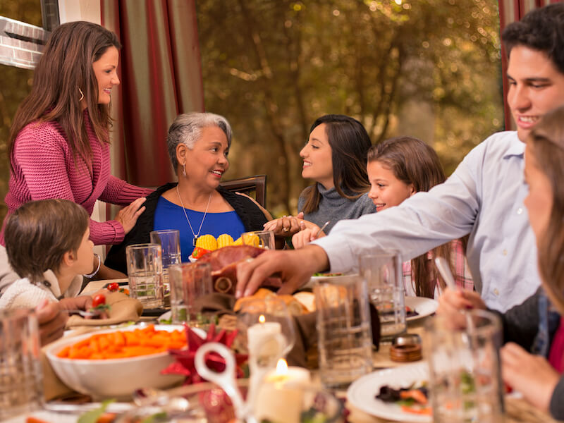 A family gathers around a dining table filled with food for a festive meal. An older woman in blue is smiling, surrounded by others of various ages engaged in conversation. The autumn setting is visible through a window.